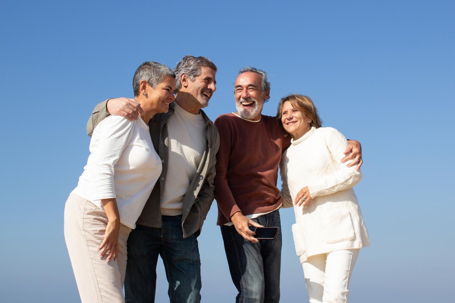 Quatro adultos maduros sorrindo abraçados ao ar livre sob céu azul.